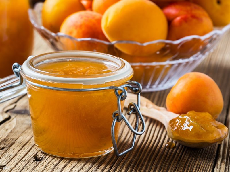 Frontal: Ein Glas Marillen-Marmelade mit Marzipan, frische Aprikosen sowie ein Löffel mit Konfitüre liegen auf einem rustikalen Holztisch.