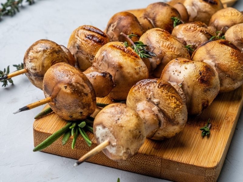 Frontal: Ein Brettchen aus Holz liegt auf einem hellem Untergrund. Auf ihm sind drei marinierte Champignon-Spieße.