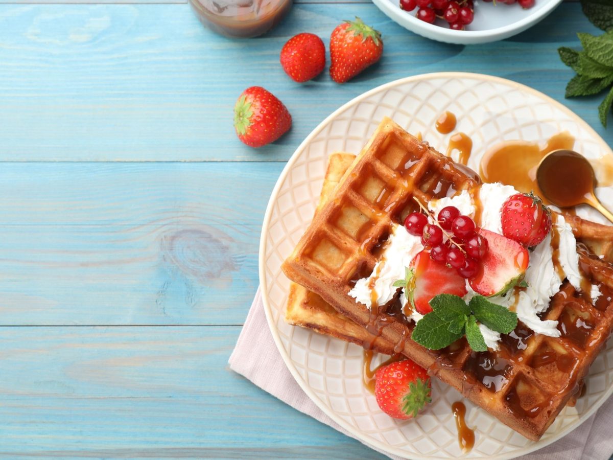 Draufsicht: Ein Teller mit süßen Mutti-Waffeln auf einem weißen Teller. Die Waffeln sind mit Eiscreme und Beeren garniert. Der Teller steht auf einem blauen Untergrund aus Holz. Drum herum liegen frische Beeren.