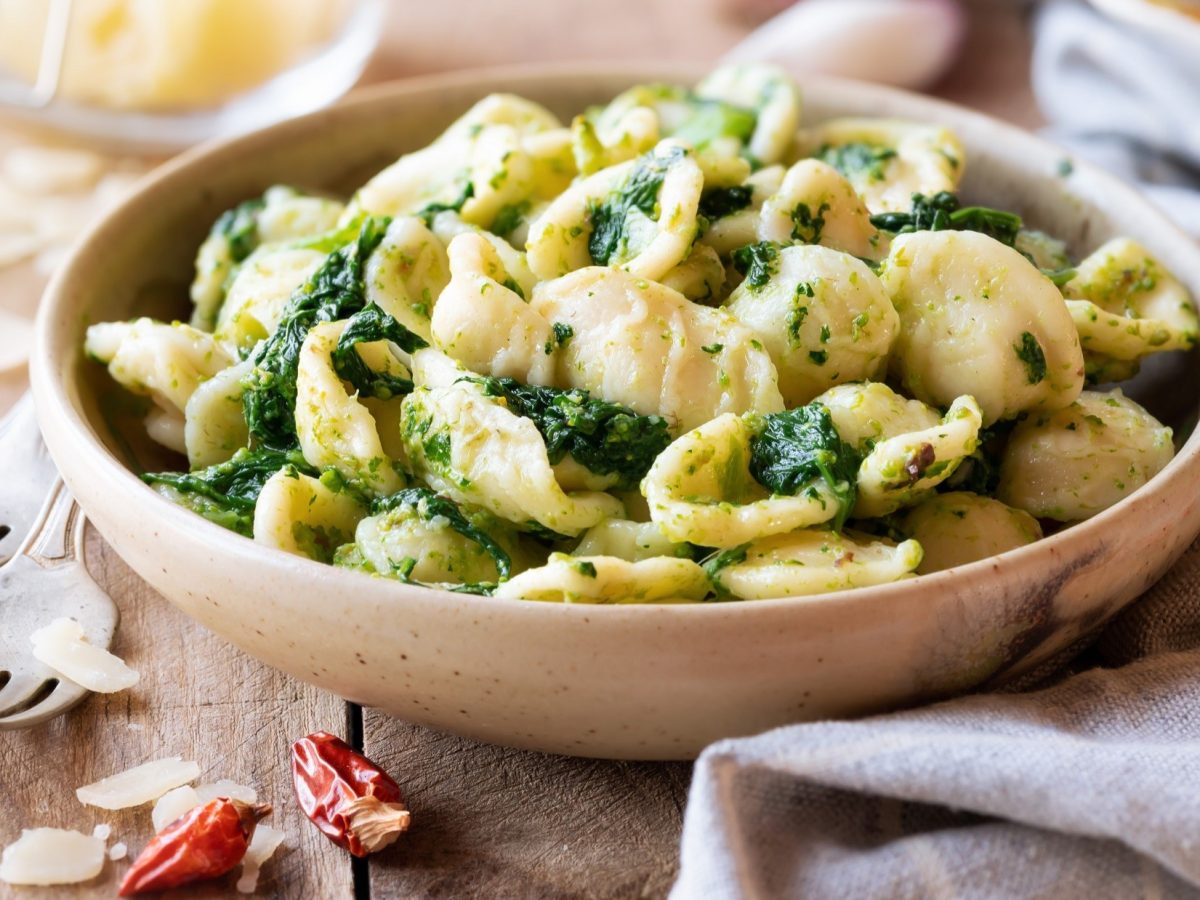 Draufsicht, schräg: Eine hellbraune Schüssel Orecchiette mit Cima di Rapa steht auf einem Holztisch. Drum herum liegen ein Küchenhandtuch, zwei Gabeln sowie eine getrocknete Chili.