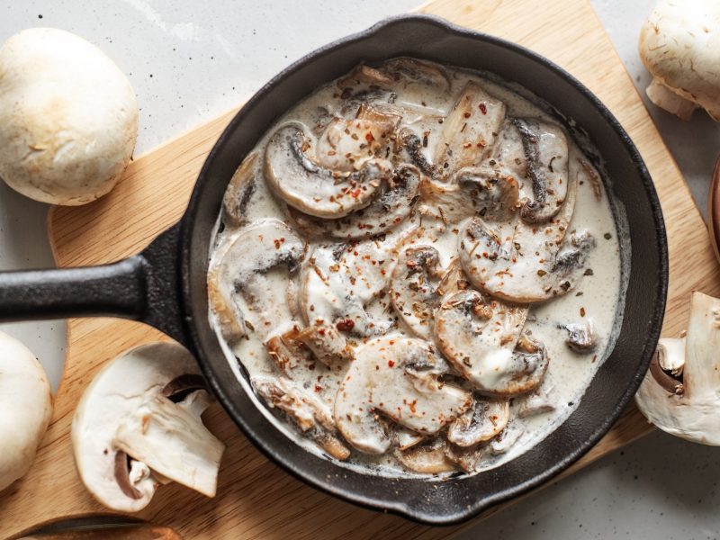 Draufsicht: Auf einem hellem Untergrund liegt eine dunkle Pfanne in der eine Pilz-Sahne-Sauce ist. Die wurde nach dem Grundrezept zubereitet. Drum herum liegen frische Champignons.