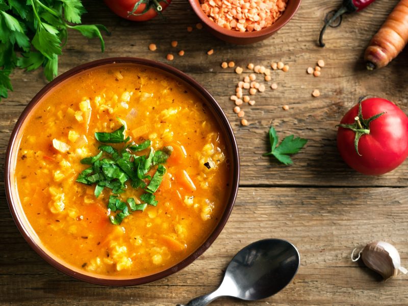 Draufsicht: Eine rustikale Unterlage aus Holz. Auf der stehe ein Teller Rote Linsensuppe mit Bulgur. Drum herum liegen frisches Gemüse und ein Suppenlöffel.