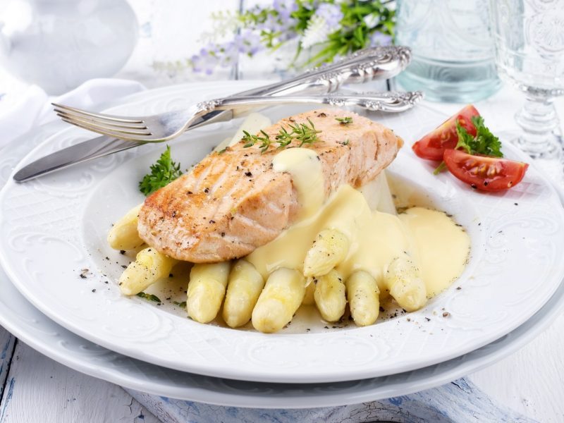 Draufsicht schräg: Ein festlich gedeckter, frühlingshafter Tisch mit einem Teller Spargel mit Lachs und Hollandaise. Am Tellerrand liegen zwei kleine Tomatenstückchen und ein massives, silbernes Besteck.