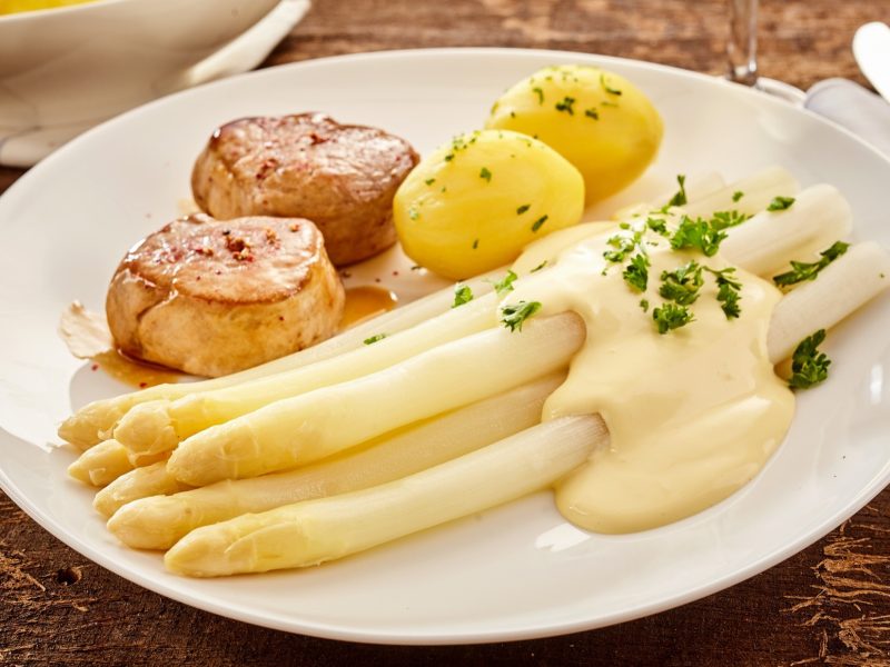 Draufsicht, schräg: Ein weißer Teller mit Spargel mit Medaillons und Sauce hollandaise liegt auf einem Untergrund auf Holz.