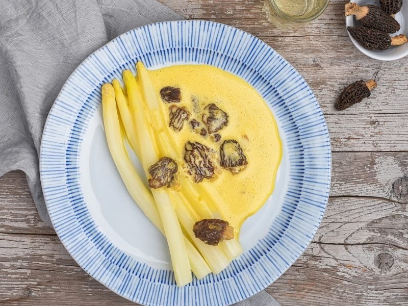 Draufsicht: Ein blau-weißer Teller Spargel mit Morchel-Cremesauce liegt auf einem Holztisch.