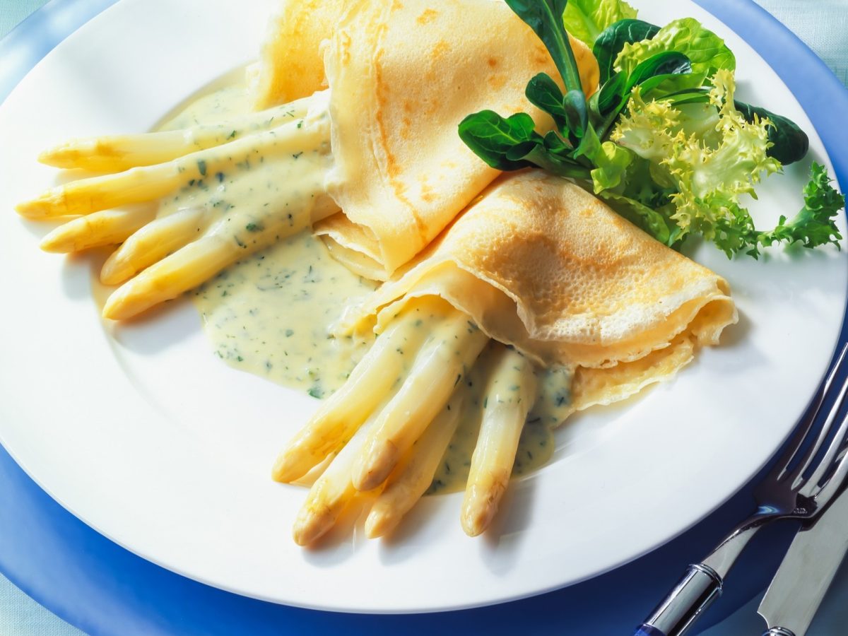 Draufsicht, Ein weißer Teller, der auf einem größeren blauen Teller liegt. Auf dem sind zwei Spargel-Pfannkuchen mit Sauce Hollandaise sowie ein paar Salatblätter. Auf dem blauen Teller liegen ein Messer und eine Gabel.