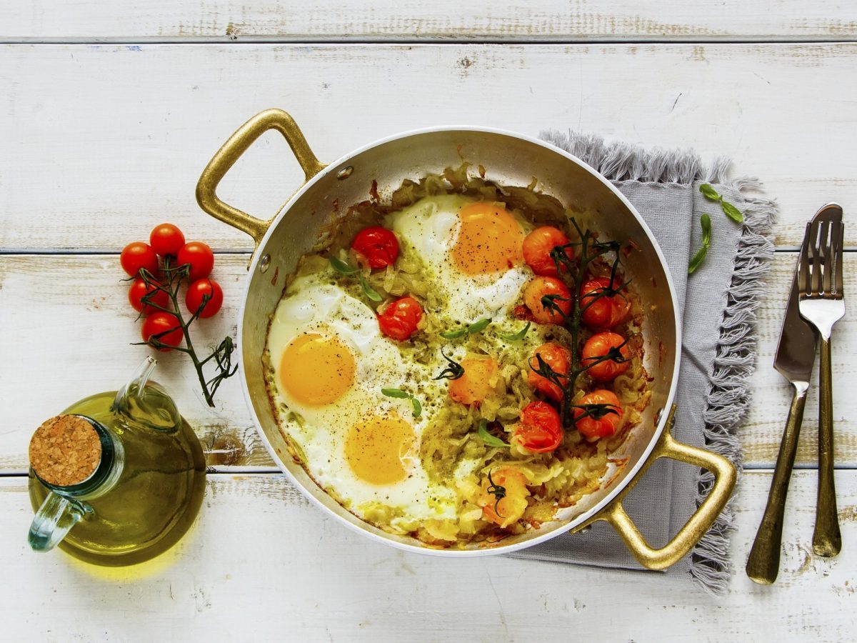 Draufsicht: Eine Pfanne Spiegeleier mit geschmorten Tomaten auf einem weißen Untergrund aus Holz. Daneben liegen Besteck, ein Küchentuch und eine Flasche mit Speiseöl.