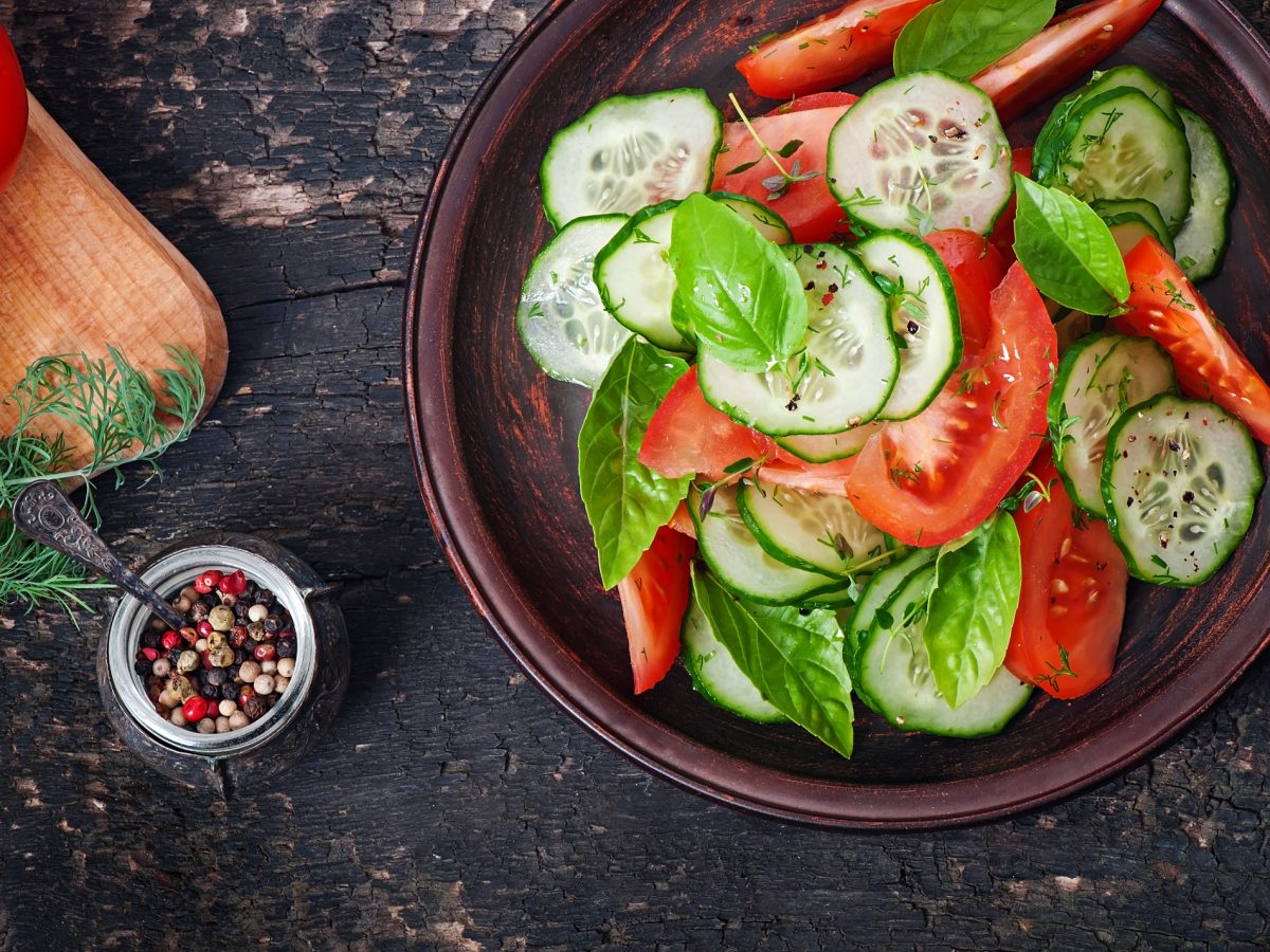 Draufsicht: Dunkler Untergrund mit einem Teller Tomaten-Gurken-Salat mit Basilikum. Daneben einen kleine Schale mit bunten Pfefferkörner und frischen Kräutern.