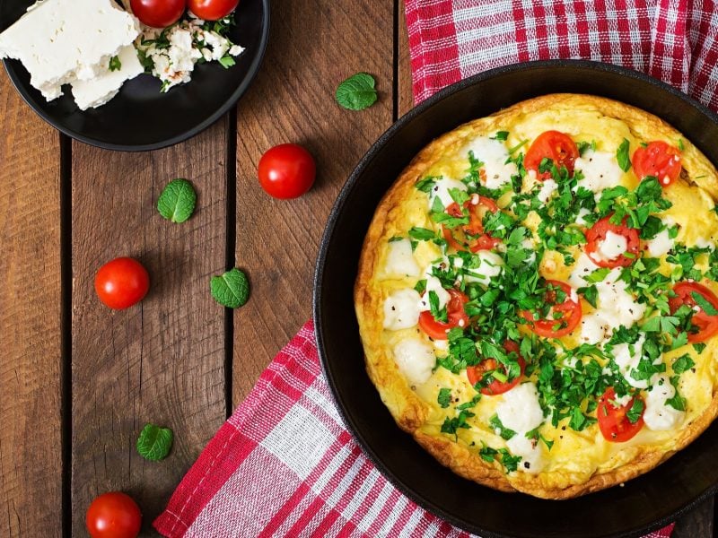 Draufsicht: Eine Pfanne mit einem Tomaten-Omelett mit Feta und Petersilie liegt auf einem rustikalen Holzuntergrund. Drum herum liegen frische Tomaten sowie ein rot-weiß-kariertes Küchentuch.