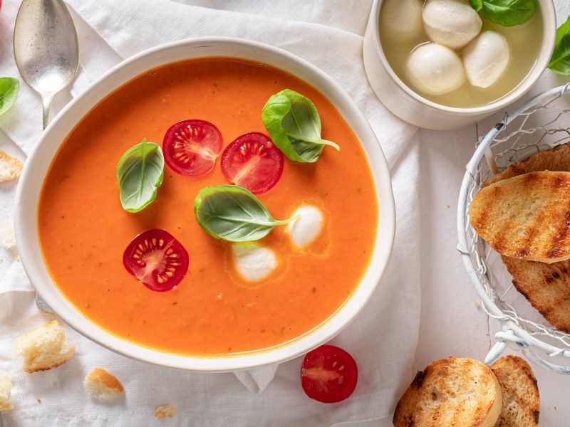 Draufsicht: Ein weißer Untergrund, auf dem eine weiße Schüssel Tomatensuppe alla Caprese steht. Drum herum gibt es eine Schalle mit Mozzarella-Kugeln, frisches Basilikum und geröstete Weissbrotscheiben.