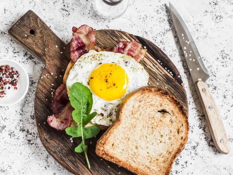 Draufsicht: Ein Uitsmijter met ham liegt auf einem Toast. Es handelt sich um ein gebratenes Spiegelei mit Bacon auf einem Toast, dass auf einem Brettchen auf einem hellen Untergrund liegt.