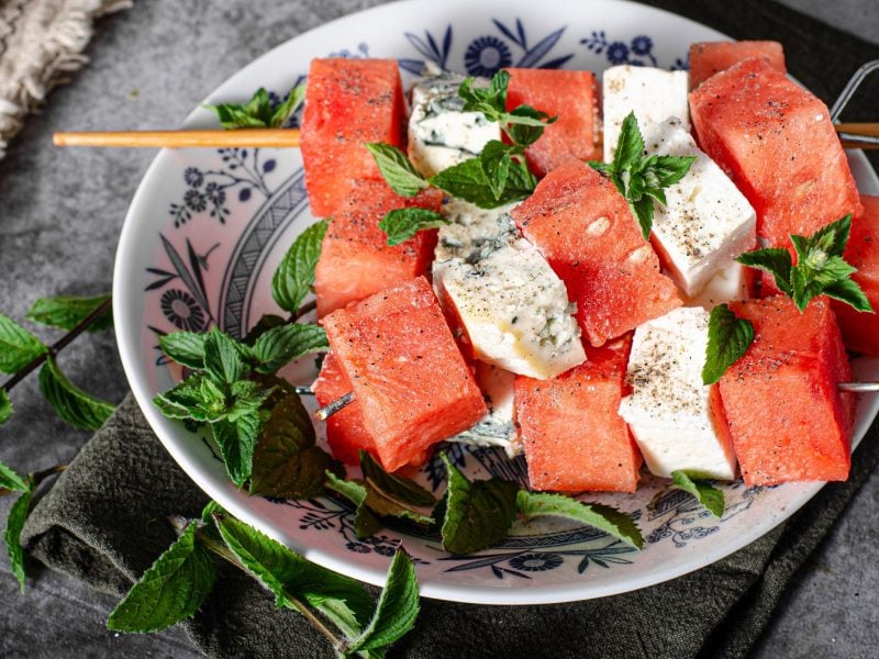 Die Melonen-Feta-Gorgonzola-Spieße auf einem weißen Teller mit blauer Verzierung und frischen Minzzweigen. Alles von oben auf einem grauen Tisch fotografiert.