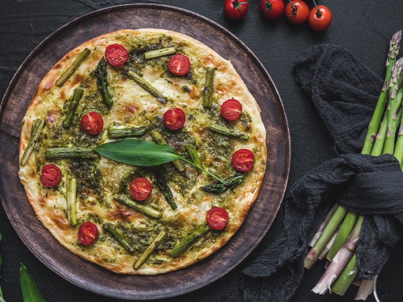 Flammkuchen mit Bärlauch-Pesto, grünem Spargel und Cherry-Tomaten vor dunklem Hintergrund, daneben frischer Spargel und Cherry-Tomaten.