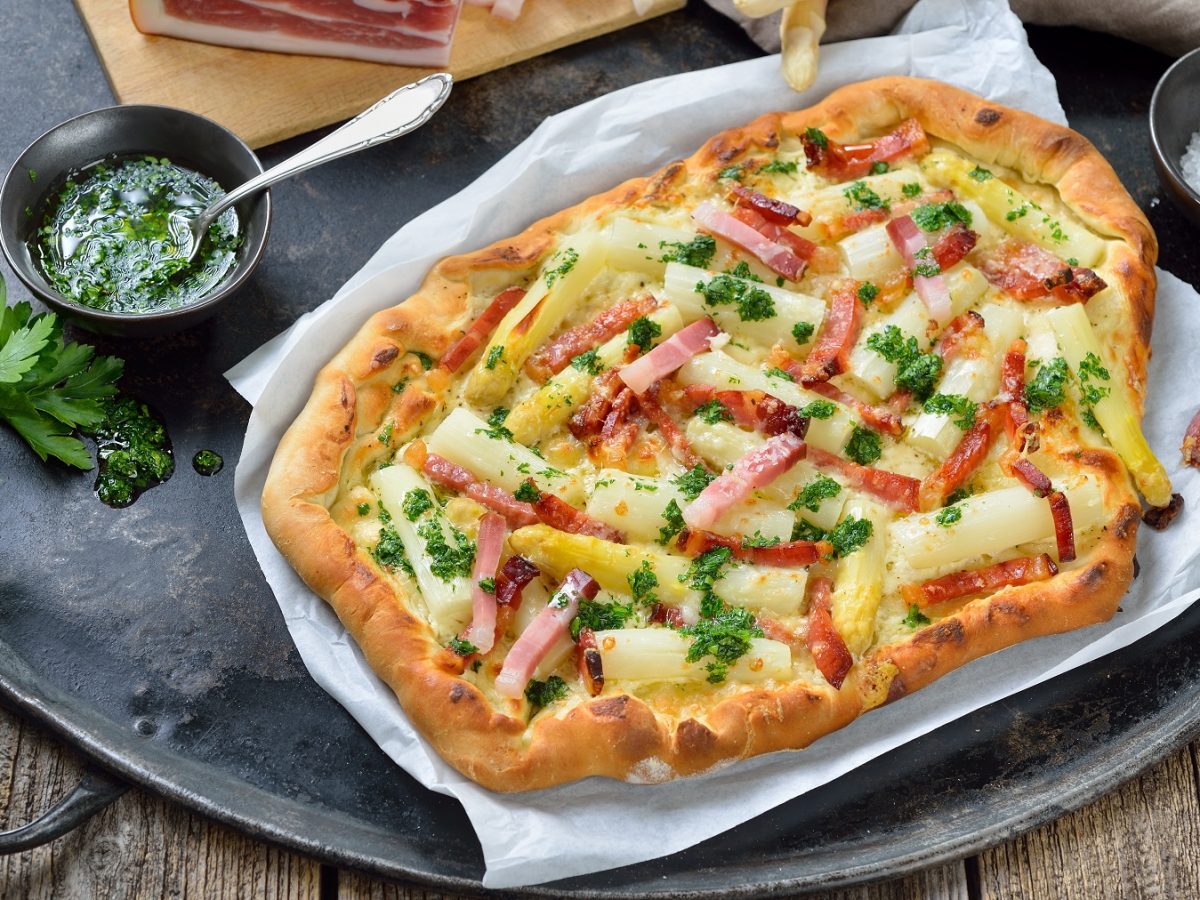 Flammkuchen mit weißem Spargel und Streifen aus Südtiroler Speck. Daneben Schüssel mit Petersilienöl. Alles auf einem schwarzen Tablett.