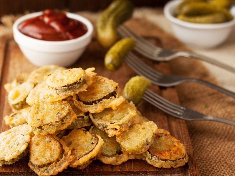 Fried Pickles im Vordergrund. Im Hintergrund Schale mit Ketchup und Essiggurken auf Gabeln.