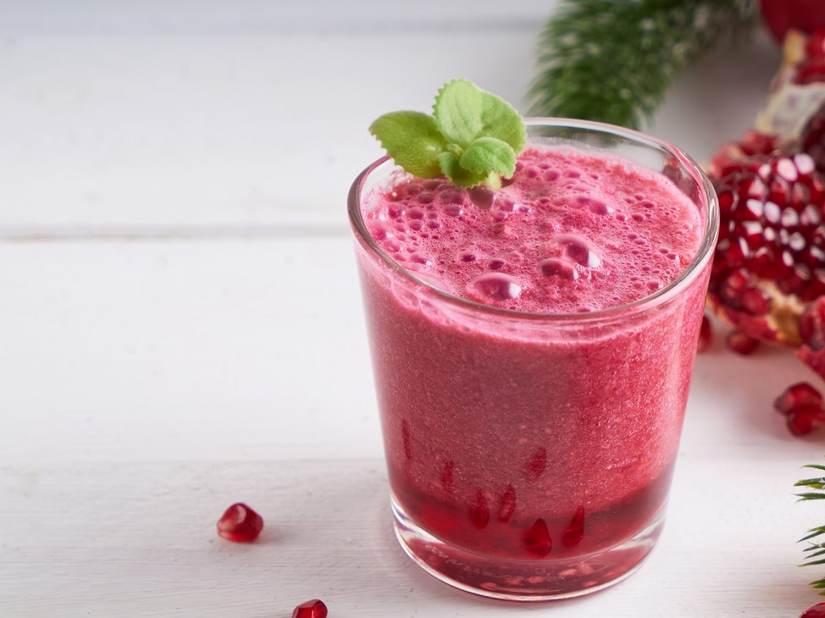 Granatapfel-Smoothie in einem Glas garnier mit frischer Minze, daneben ein angeschnittener Granatapfel.