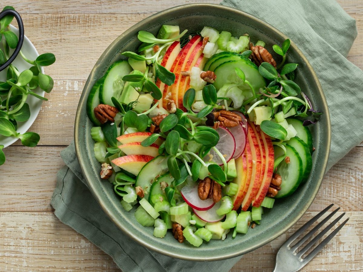 Ein dunkelgrüner Teller mit dem Gurkensalat auf einem Holztisch direkt von oben fotografiert. Darunte reien hellgrüne Serviette und daneben ein paar Sprossen.