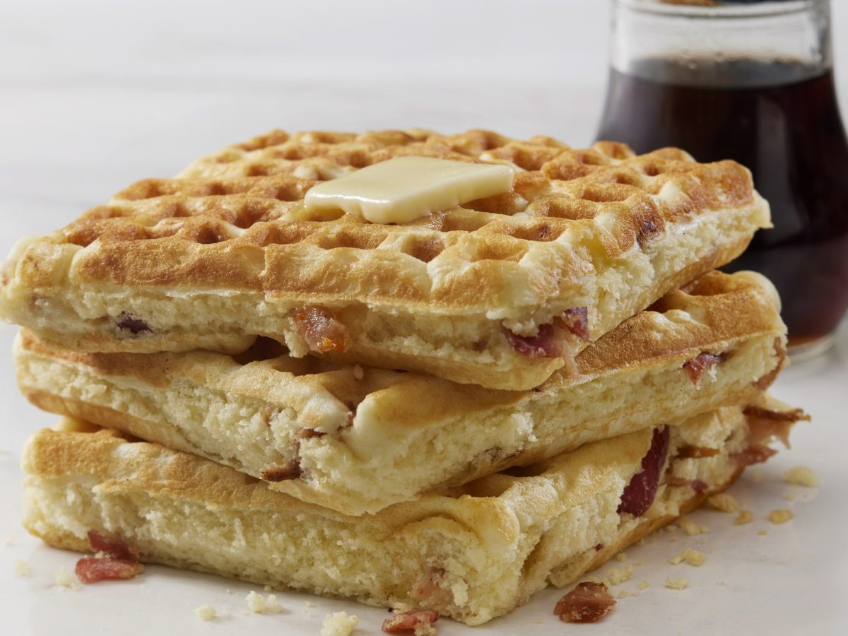 Frisch gebackene herzhafte Waffeln mit schmelzender Butter und Speckstücken, Ahornsirup im Hintergrund.