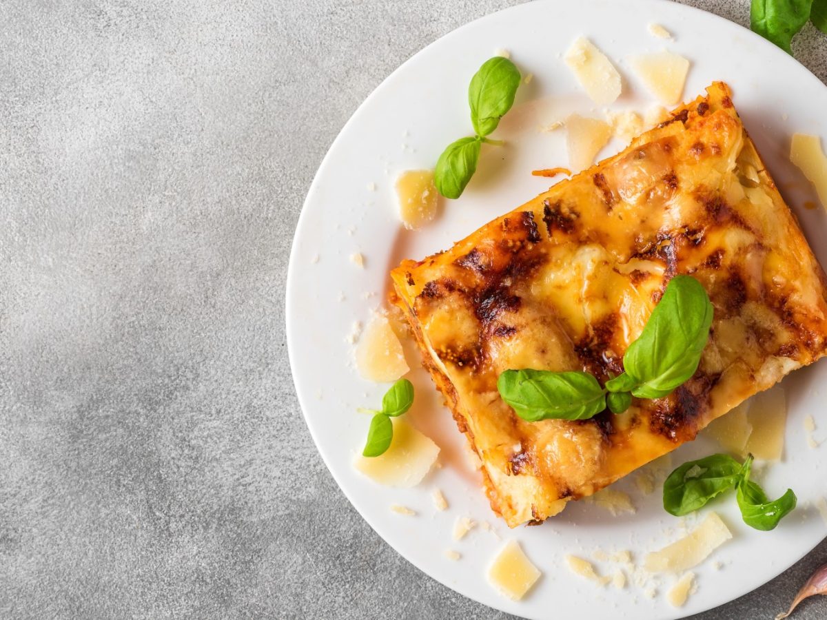 Ein Stück vegane Hummus-Lasagne auf einem weißen Teller. Darauf veganer Parmesan verteilt. Der Hintergrund ist grau.