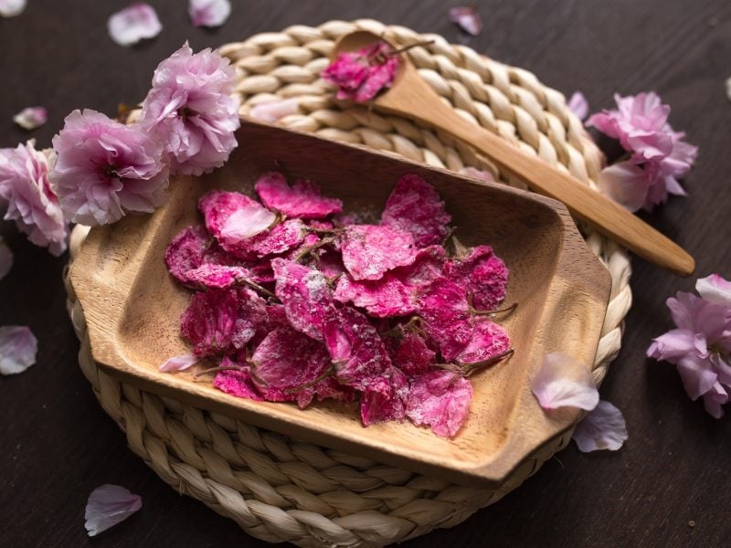 Ein Holzkörbchen mit den gesalzenen Kirschblüten und drumherum frische Kirschblüten. Alles auf einem dunklen Holztisch direkt von oben fotografiert.