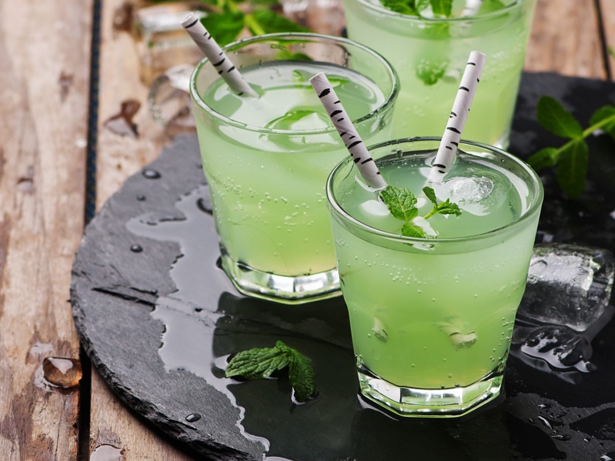 Drei Gläser mit dem grünen Drink auf einer schwarzen Schiefertafel und rustikalem Holztisch. Alles von der Seite fotografiert.