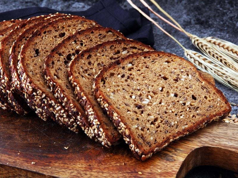 Das angeschnittene Brot auf einem Holzschneidebrett und dunkelblauem Untergrund. Dahinter frisches Getreide. Alles von schräg oben fotografiert.