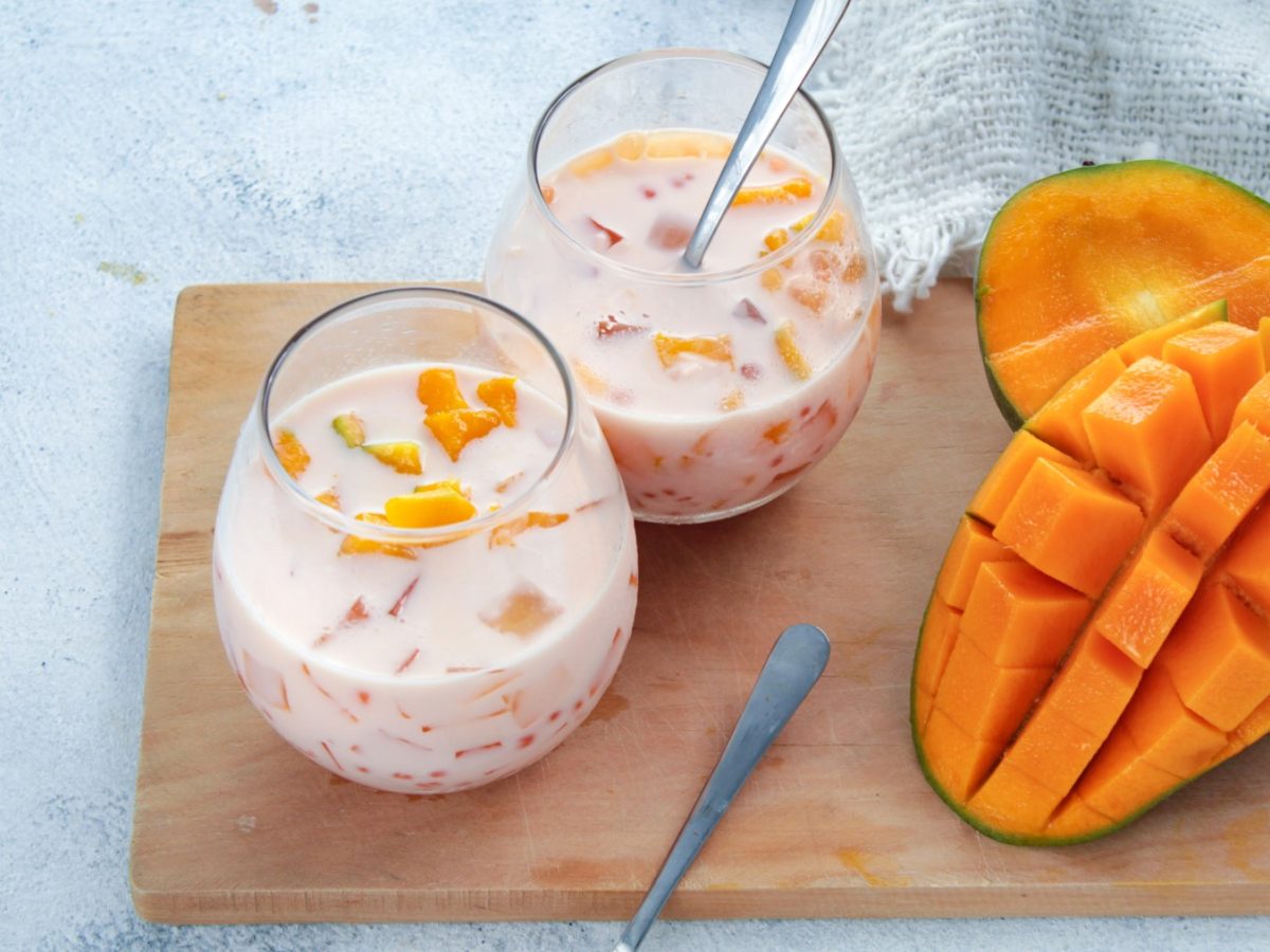 Mango Sago in Gläsern auf einem hölzernen Brett. Rechts auf dem Foto halbe Mangos und der Hintergrund ist hellgrau.