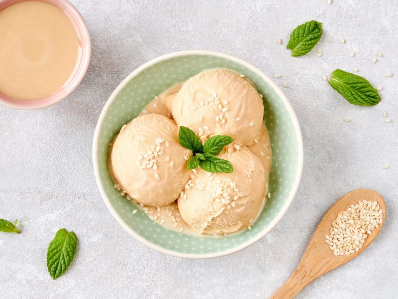 Drei Kugeln Milchmädchen-Eis mit Tahini in einer Keramikschüssel, mit Sesamsamen. Daneben eine Schüssel mit Tahini.