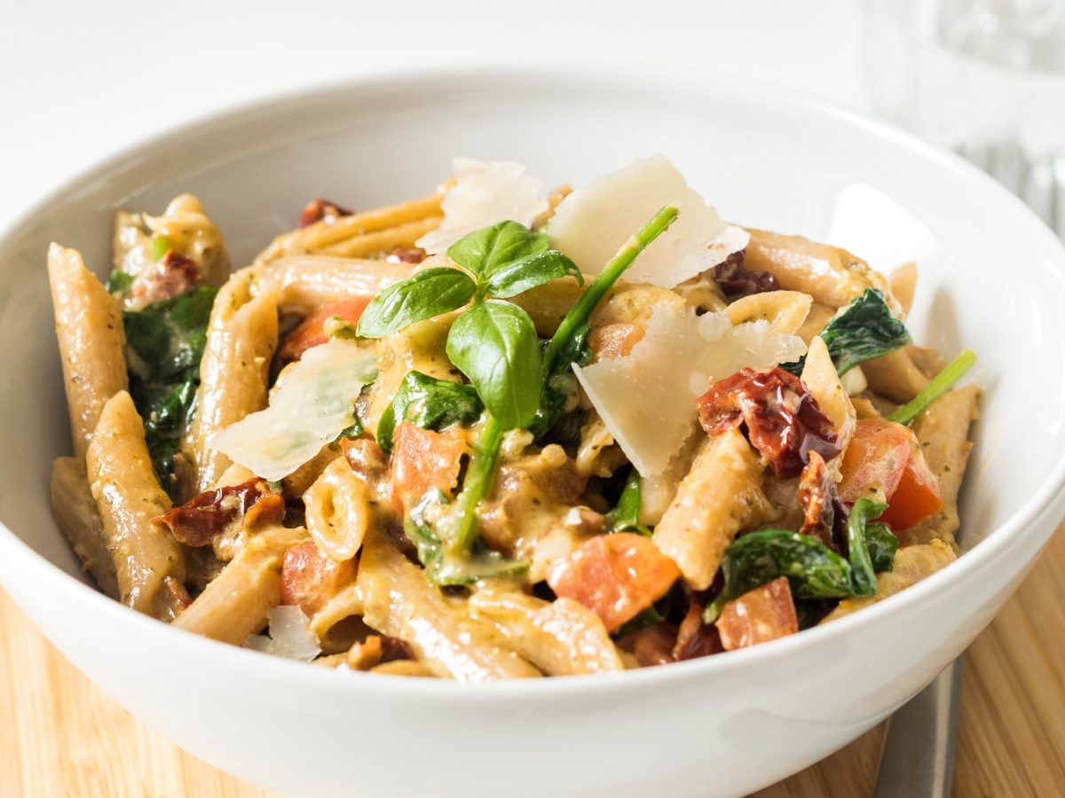 Eine weiße Schale mit dem Nudelsalat auf einem runden Holzbrett und hellem Hintergrund. Alles von leicht schräg oben fotografiert.