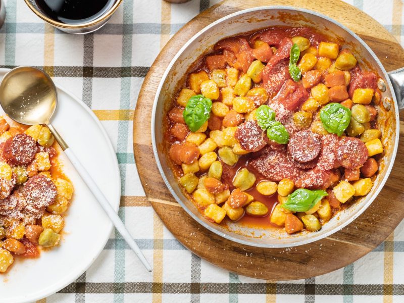 Eine Pfanne und ein weißer Teller mit den Gnocchi auf einem Tisch mit blau-gelb-weiß gestreifter Tischdecke. Danebn noch Salz- und Pfefferstreuer sowie ein Glas mit Rotwein. Alles direkt von oben fotografiert.