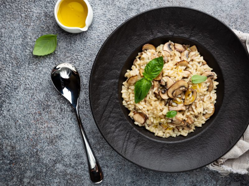 Ein schwarzer Teller mit der One-Pot-Pasta auf grauem Tisch mit Löffel und kleinem weißen Öl-Kännchen. Alles direkt von oben fotografiert.