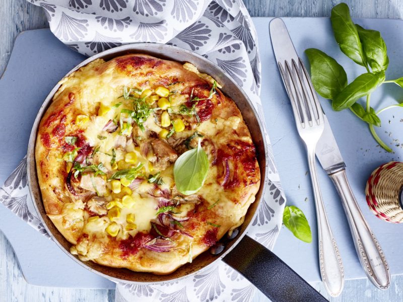 Draufsicht: Pfannen-Pizza mit frischem Basilikum auf in der Pfanne mit hellblauem Untergrund.