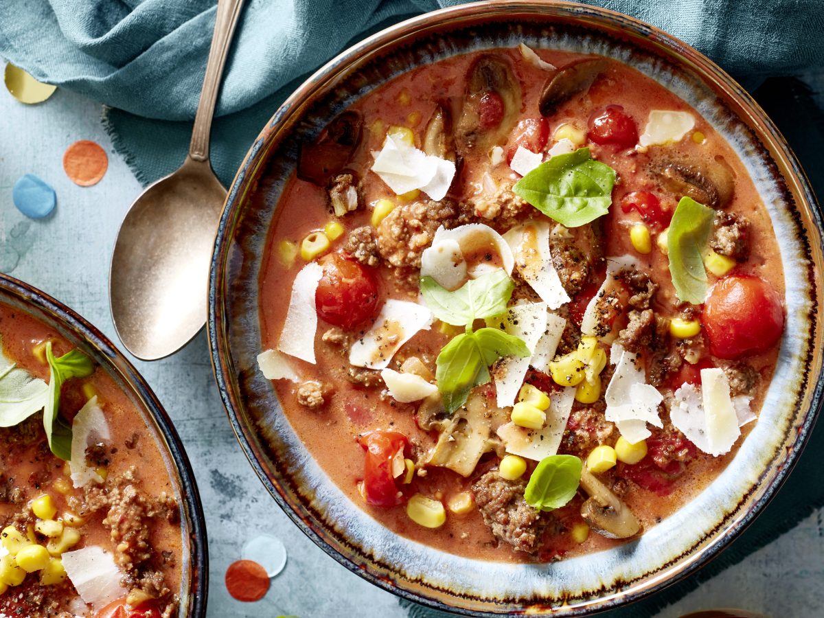 Pizzasuppe mit Hackfleisch, Mais und Gemüse, bestreut mit Käse, auf hellem Steinuntergrund.