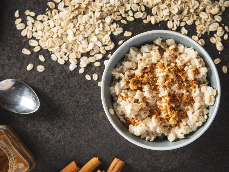 Porridge auf Basis eines Grundrezepts in einer weißen Schüssel mit Zimt. Daneben Löffel, Haferflocken und Zimtstangen.