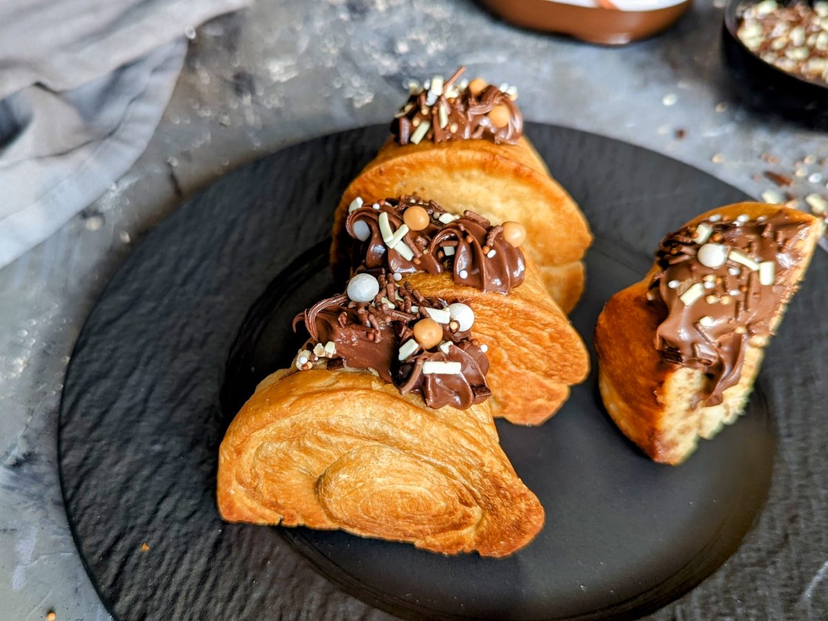 Einige Croissant-Rolls mit Nutella gefüllt auf einem schwarzen Teller und grauem Steinuntergrund. Im Hintergrund eine graue Tischdecke und ein schwarzes Schälchen mit der Schoko-Deko.