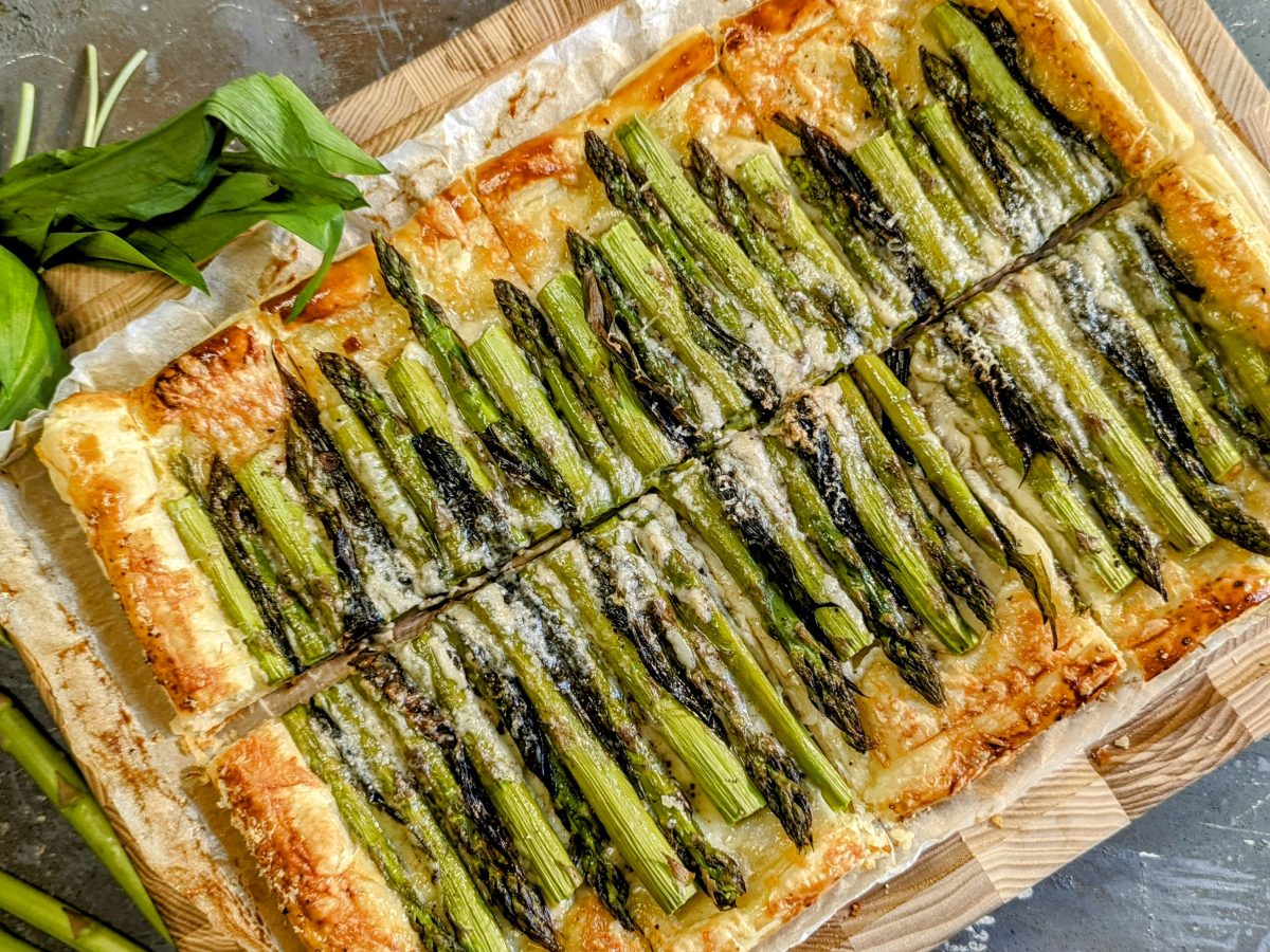 Die eckige Tarte mit Spargel auf einem Holzbrett und grauem Untergrund direkt von oben fotografiert. Links daneben noch einige Kräuter und Spargel.