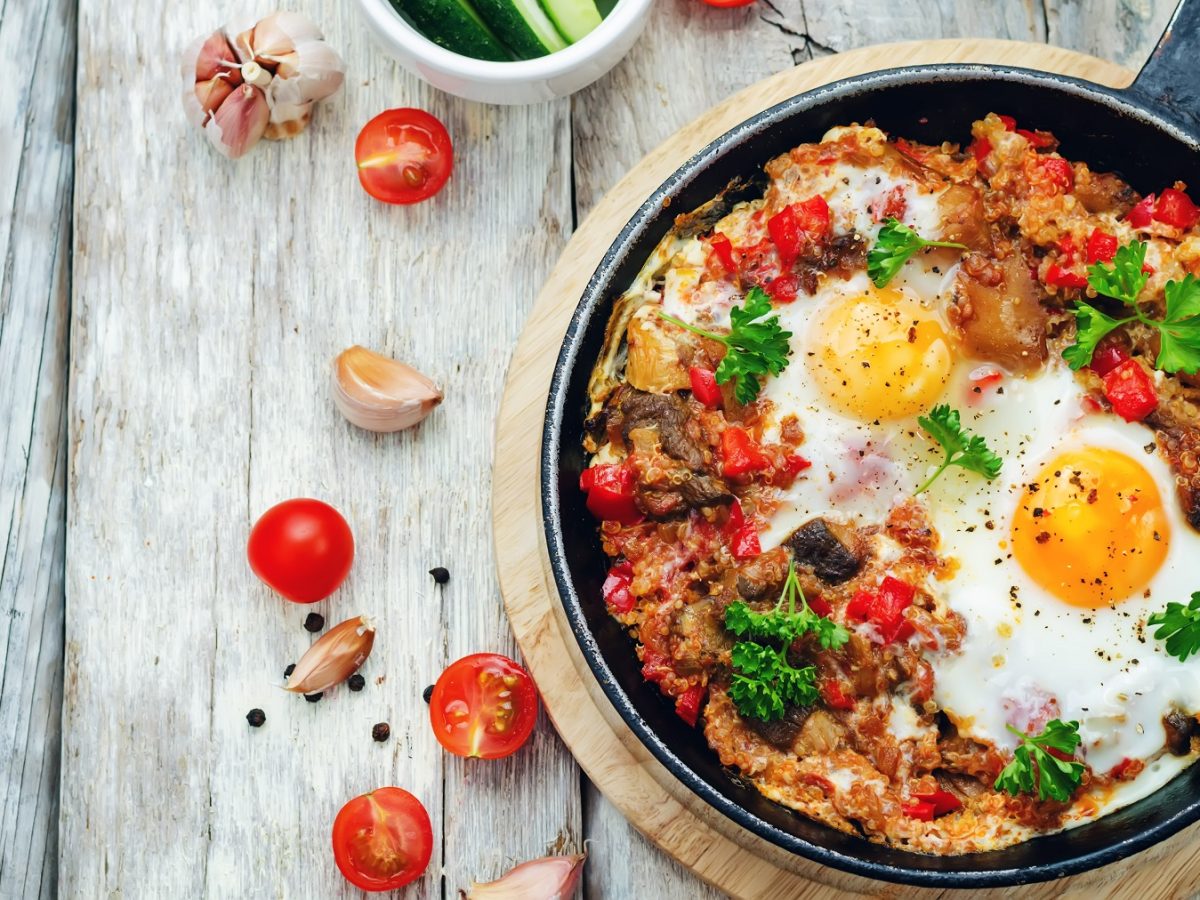 Quinoa-Pfanne mit Gemüse und Eiern in einer Pfanne. Daneben angeschnittene Tomaten und Knoblauchzehen.