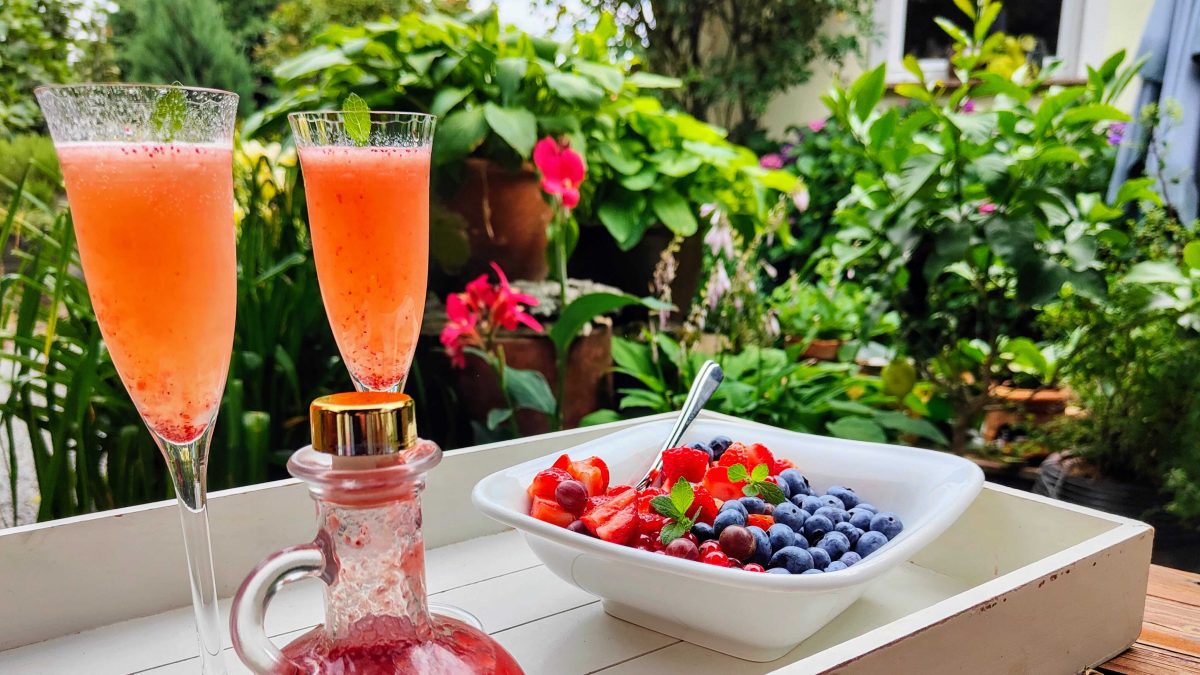 Zwei Gläser mit einem Retro Strawberry Spritz in einem grünem Garten. Davor steht eine Schale voll Beerenfrüchte.