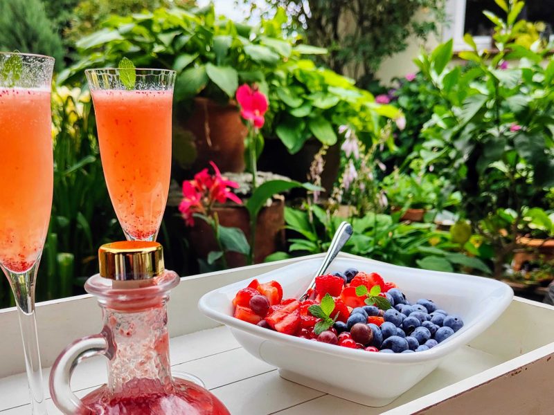 Zwei Gläser mit einem Retro Strawberry Spritz in einem grünem Garten. Davor steht eine Schale voll Beerenfrüchte.