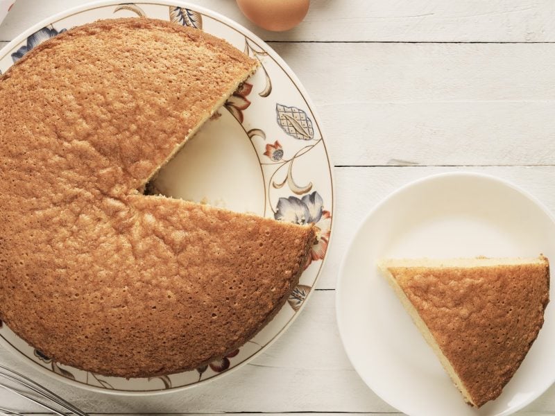 Ein Stück Rührkuchen aus Rührteig auf weißem Teller, neben ganzen Kuchen auf Holztisch.