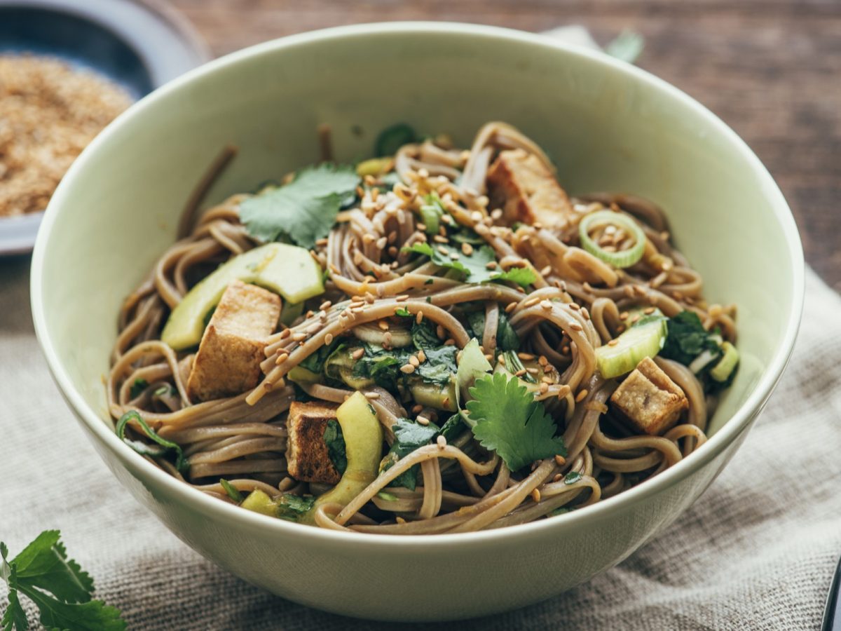 Eine hellgrüne, tiefe Schale mit dem Soba-Salat von schräg oben auf einem Holztisch mit Leinentuch fotografiert.