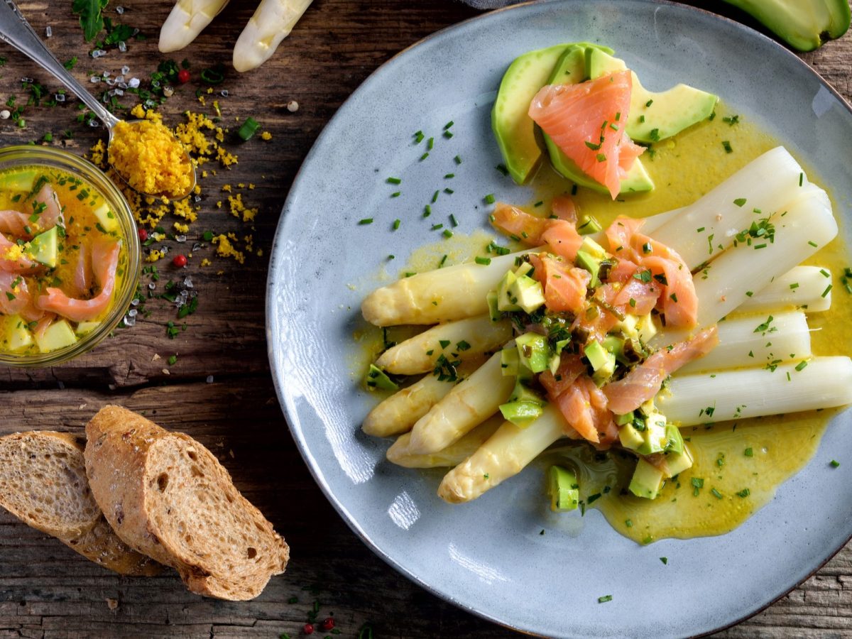 Spargel mit Lachs, Avocado und Zitronensauce auf einem Teller, daneben frisches Brot und eine Schale mit den Hauptkomponenten.