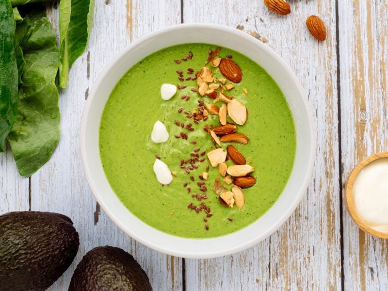 Spargel-Smoothie-Bowl in einer weißen Schüssel auf weißem Hintergrund. Links unten Avocado, links oben Spinat.