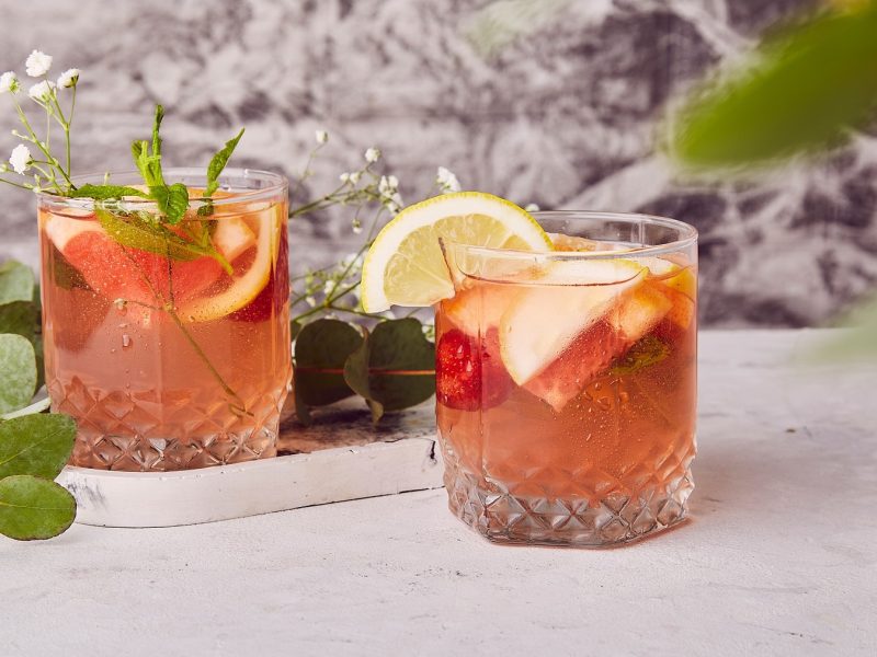 Zwei Gläser mit dem Spring Negroni auf einem hellen Tisch mit grauem Hintergrund. Daneben und dazwischen grüne Pflanzenblätter.