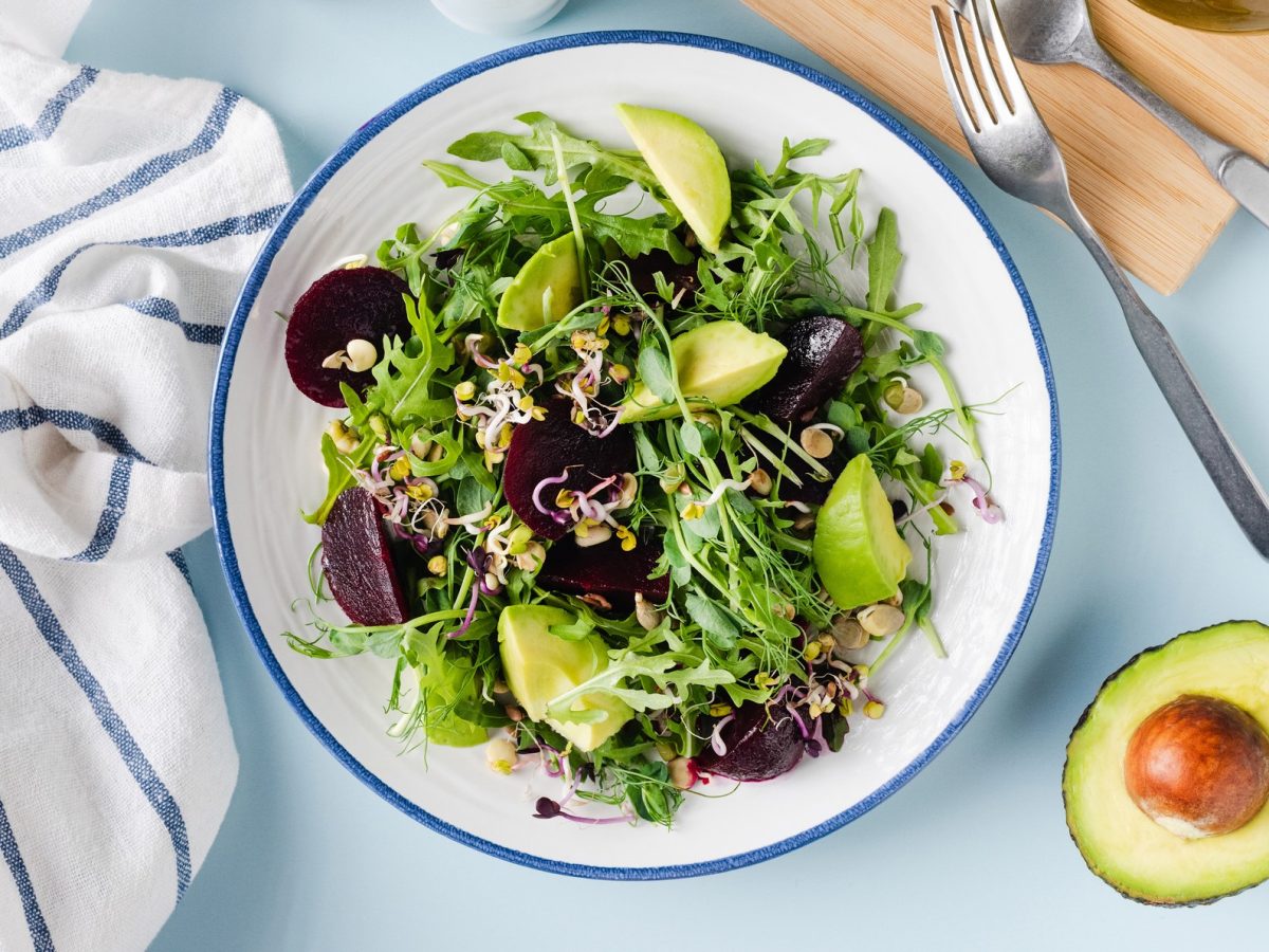 Sprossensalat mit Roter Bete und Avocado auf einem hellen Teller. Daneben eine angeschnittene Avocado mit Kern.