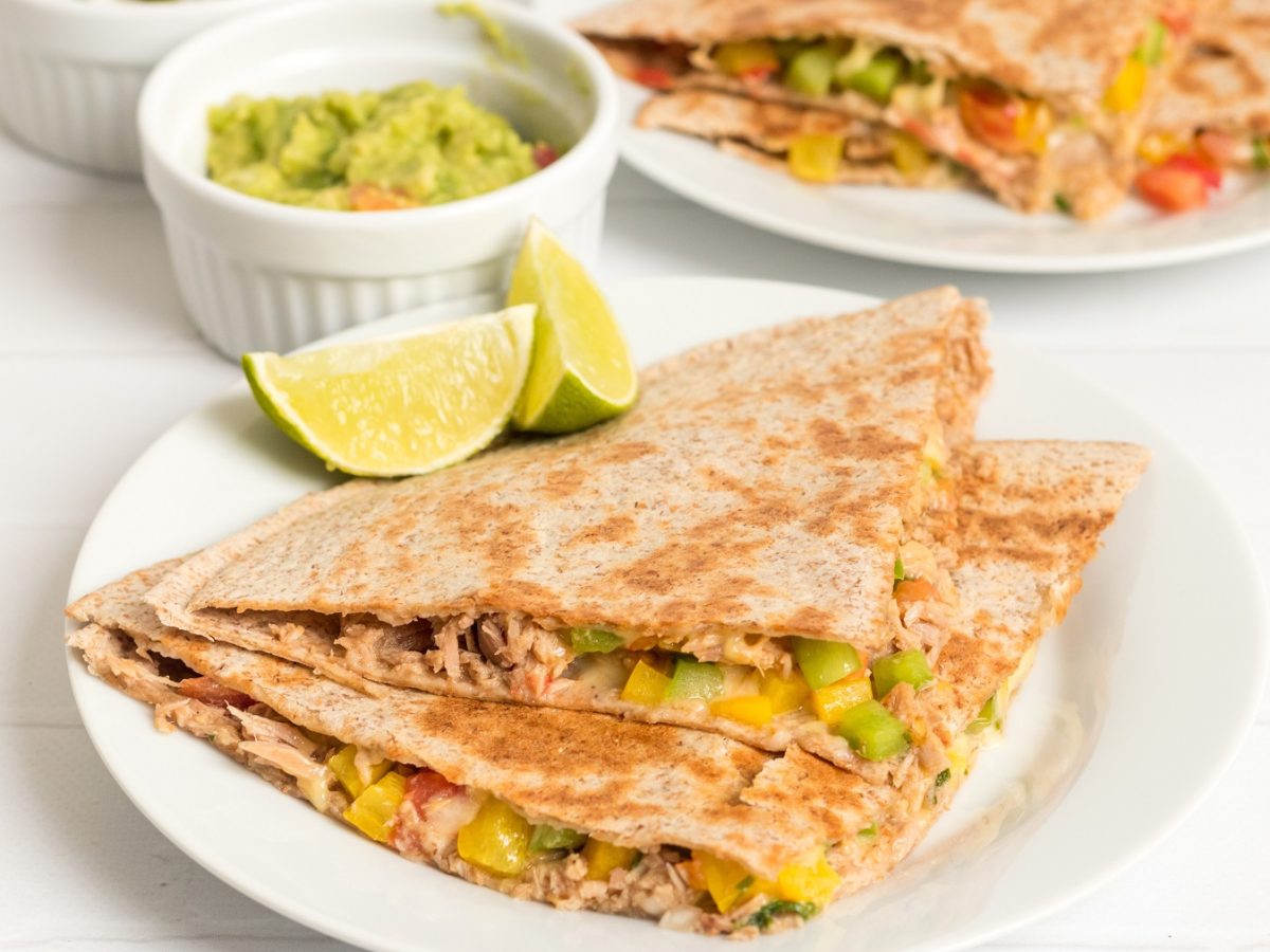 Jeweils 2 Ecken der Quesadilla auf zwei weißen Tellern mit Limettenspalten und einem weißen Schälchen Guacamole. Alles steht auf einem weißen Tisch und ist von der Seite fotografiert.