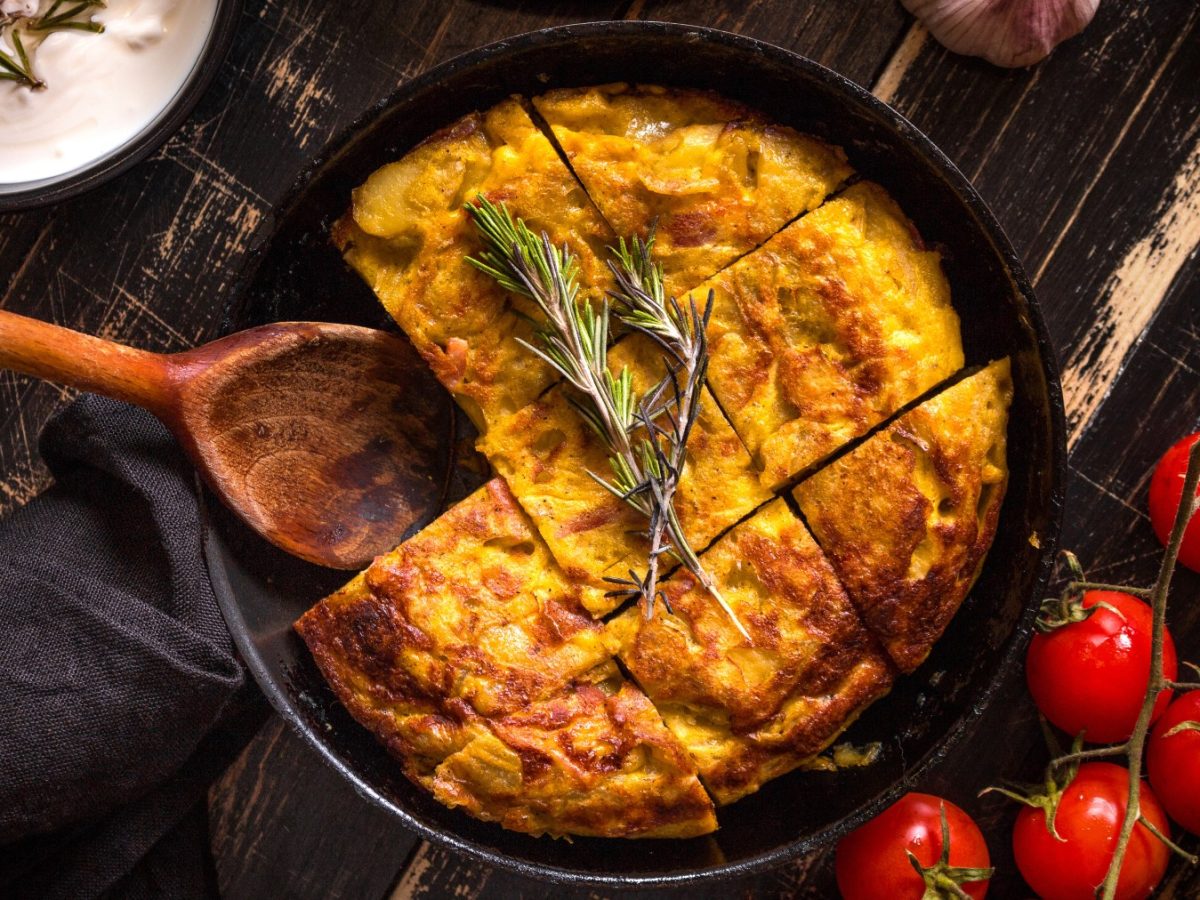 Tortilla de Patatas in einem schwarzen Teller in Stücke geschnitten auf dunklem Hintergrund. Rechts Tomaten und Besteck, links ein Schüsselchen mit einem Dip und ein Löffel.