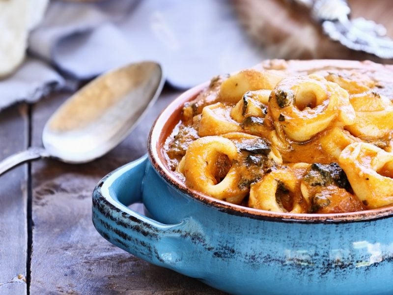 Eine blaue Auflaufform mit den überbackenen Tortellini. Alles auf einem Holztisch von der Seite fotografiert.