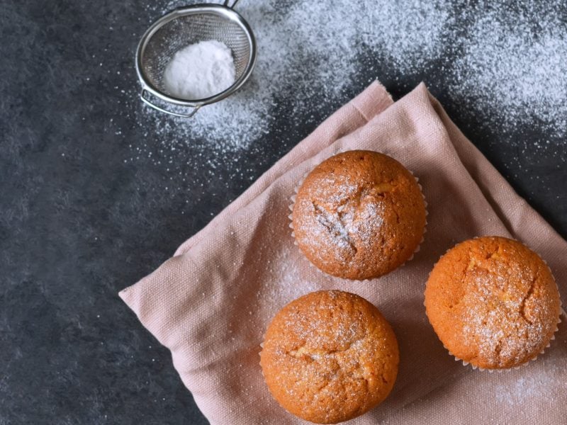 Vegane Muffins, mit Puderzucker bestäubt auf einem Küchentuch vor grauem Untergrund.