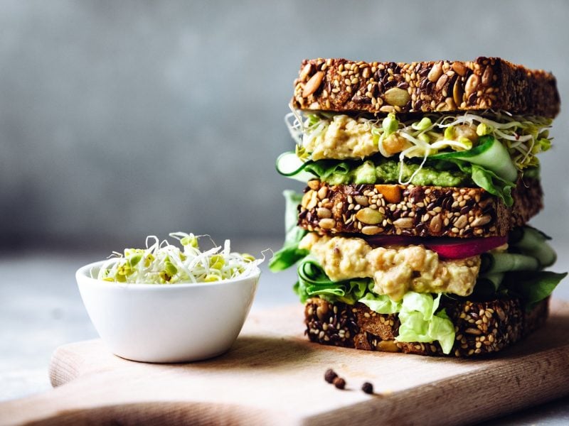 Veganes Sandwich mit Kichererbsensalat und Sprossen auf einem Brett.